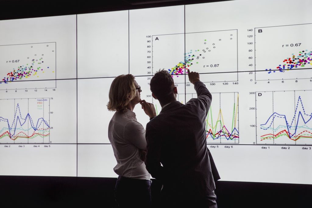Two people pointing at graphs on a large screen.