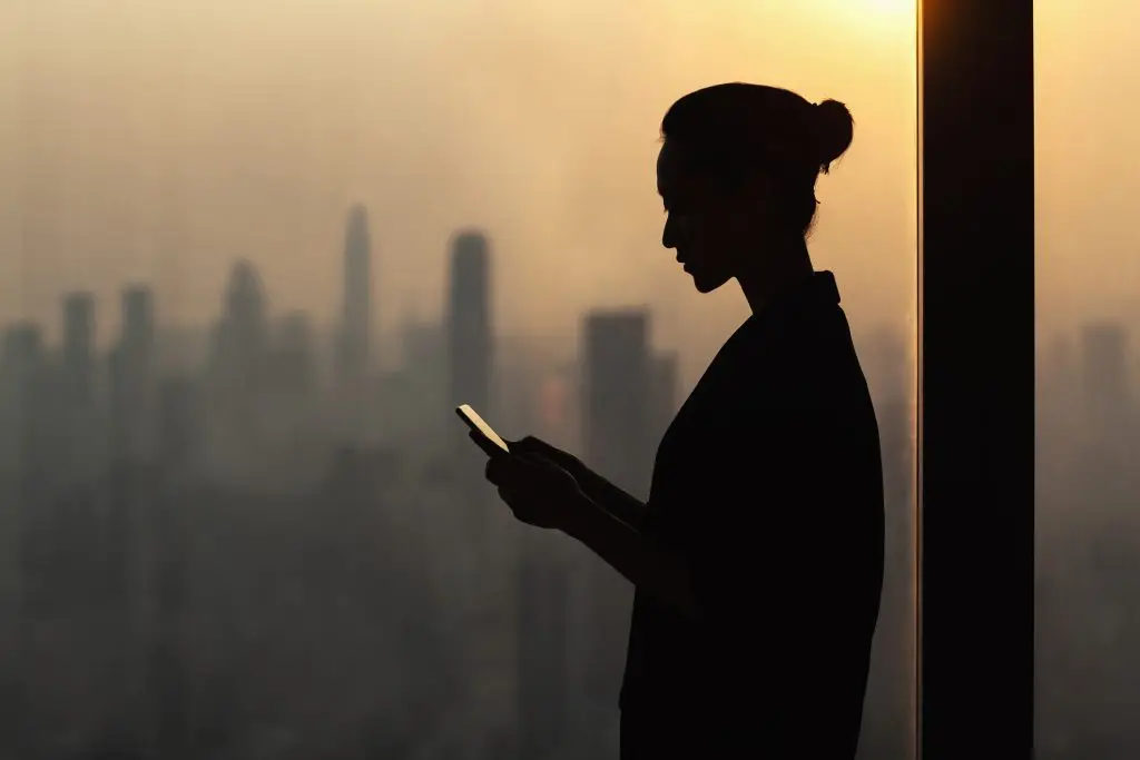 A silhouette of a woman using a cell phone in front of a city.