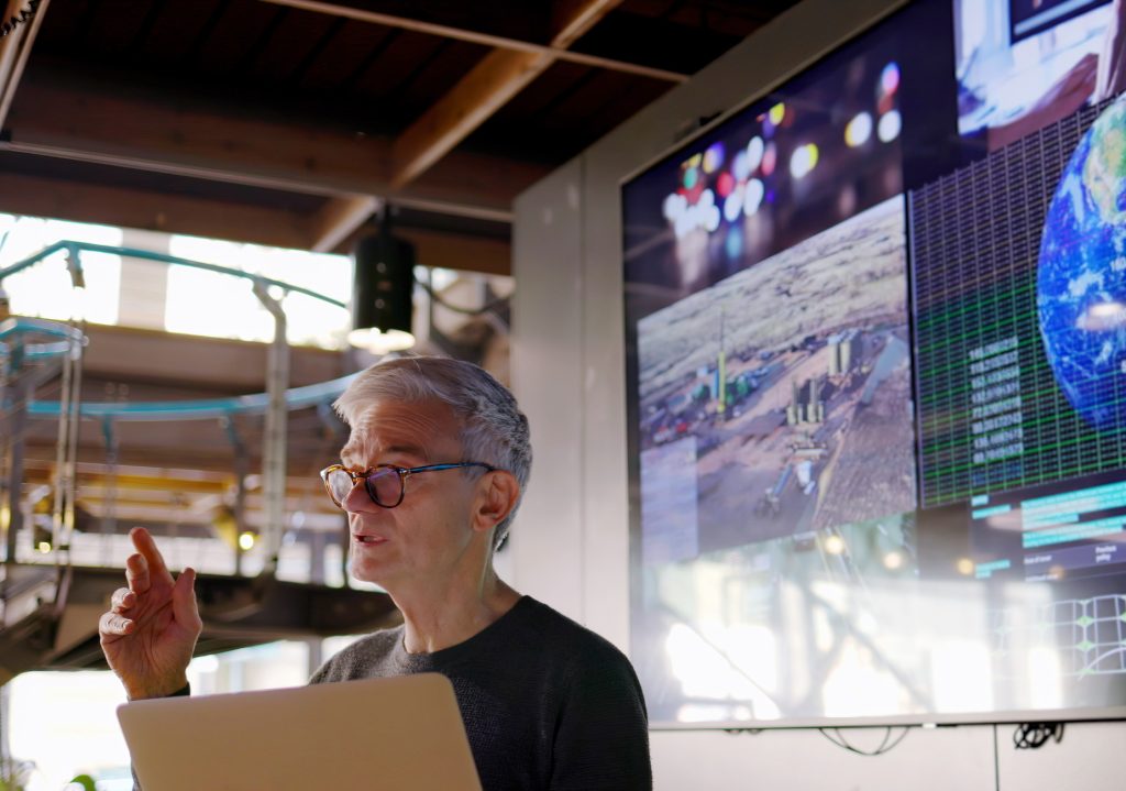 A man in glasses is giving a presentation in front of a large screen.