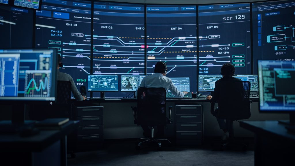 Three people work at a control room with multiple computer monitors and a large digital display showing complex data and graphs.