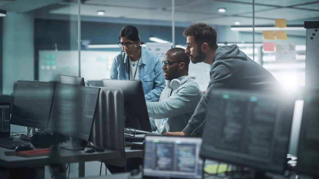 Three people collaborate in an office with computer screens displaying code. The group appears focused, engaged in problem-solving. The setting is modern with glass walls and workstations.