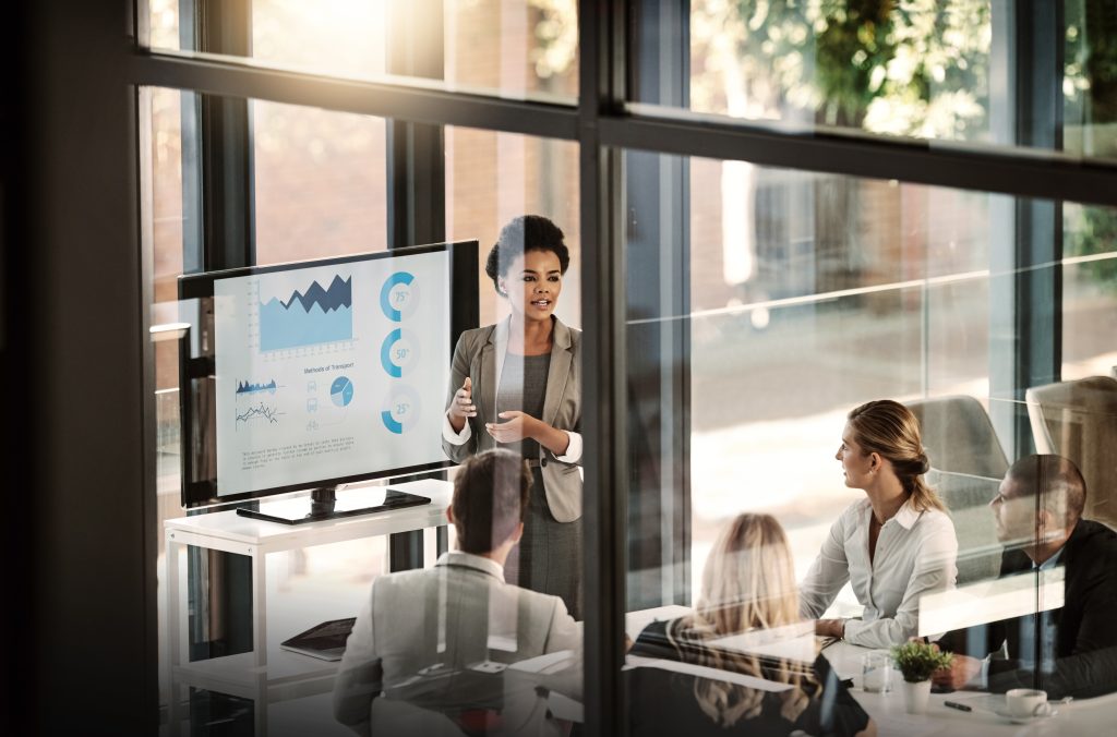 A woman presents charts on a screen to four colleagues seated around a table in a glass-walled office.
