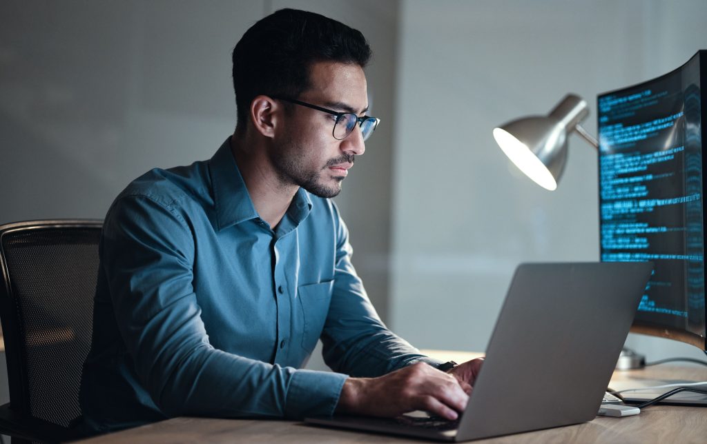 Man wearing glasses sits at a desk working on a laptop, with code visible on a large monitor and a desk lamp providing light.