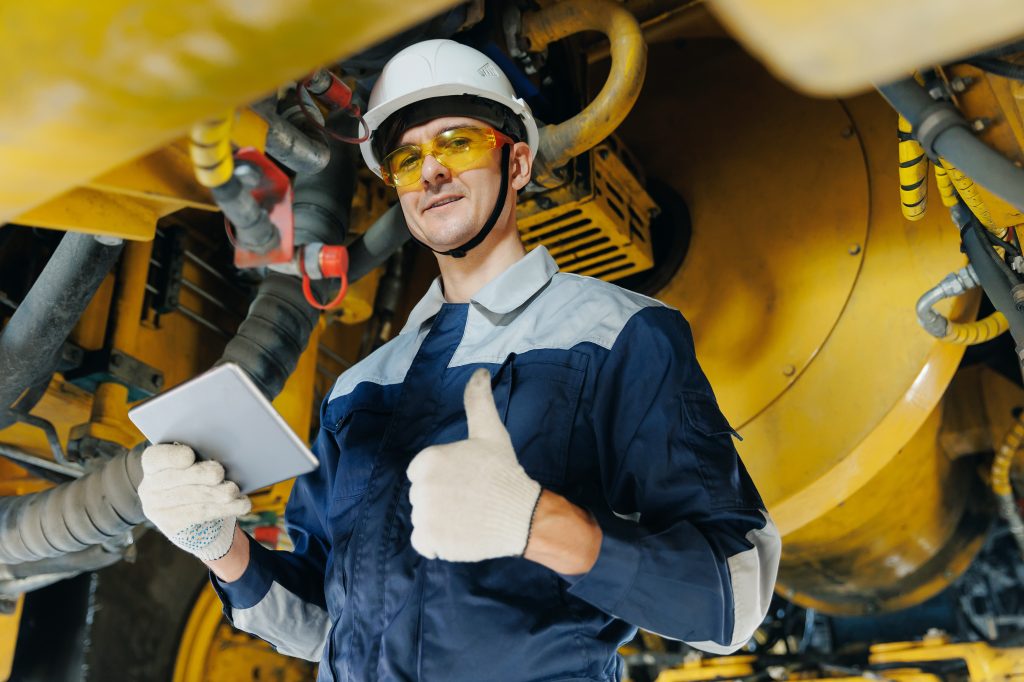 A worker in safety gear holding a tablet gives a thumbs up while standing under heavy machinery in an industrial setting.
