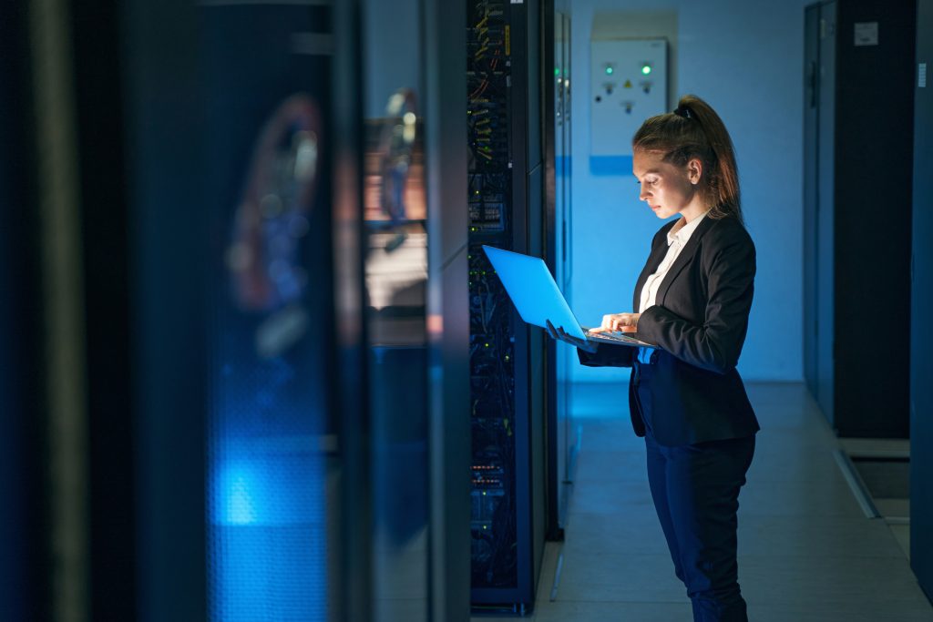 A woman in business attire uses a laptop while standing in a server room, surrounded by racks of computer equipment and blue lighting.