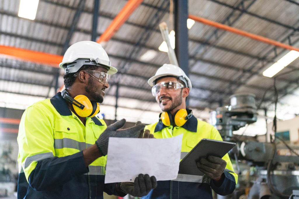 Two factory workers in safety gear discuss a document and a tablet inside an industrial facility.