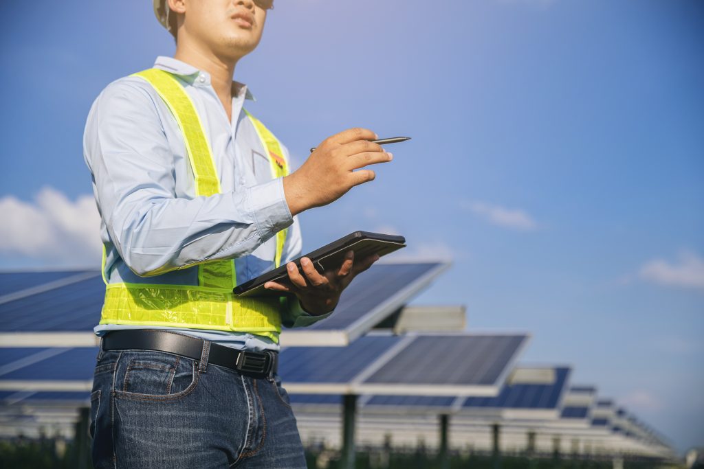 Person in safety vest and helmet holds tablet and pen, standing in front of solar panels under a clear sky.