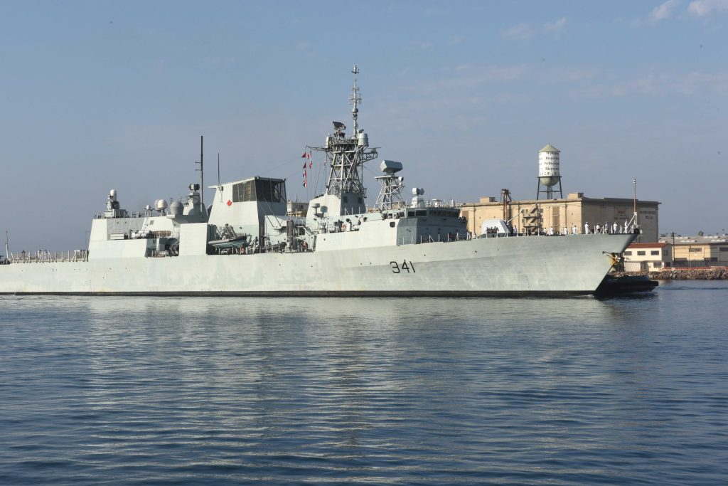 A naval ship marked with the number 341 is docked near a building with a water tower, under a clear sky.