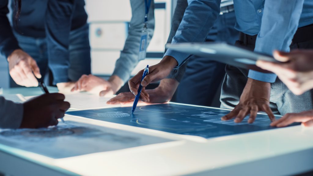 Several people stand around a touchscreen table, examining and pointing at technical blueprints or plans displayed on the screen.