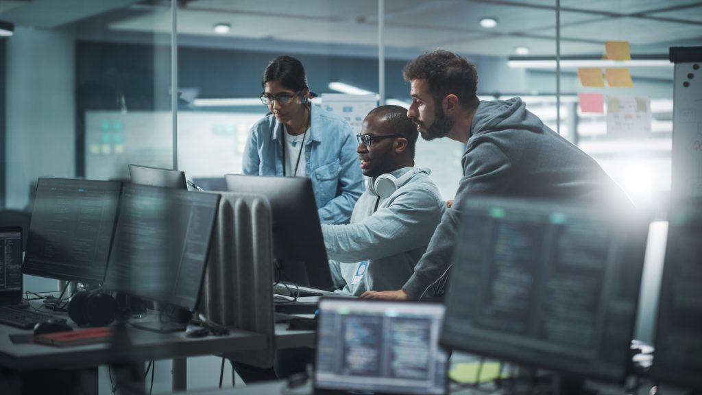 Three people work together at computer monitors in a modern office, viewing code on screens in a collaborative tech environment.