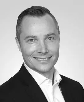 A man in a suit smiles at the camera against a plain background. The image is in black and white.