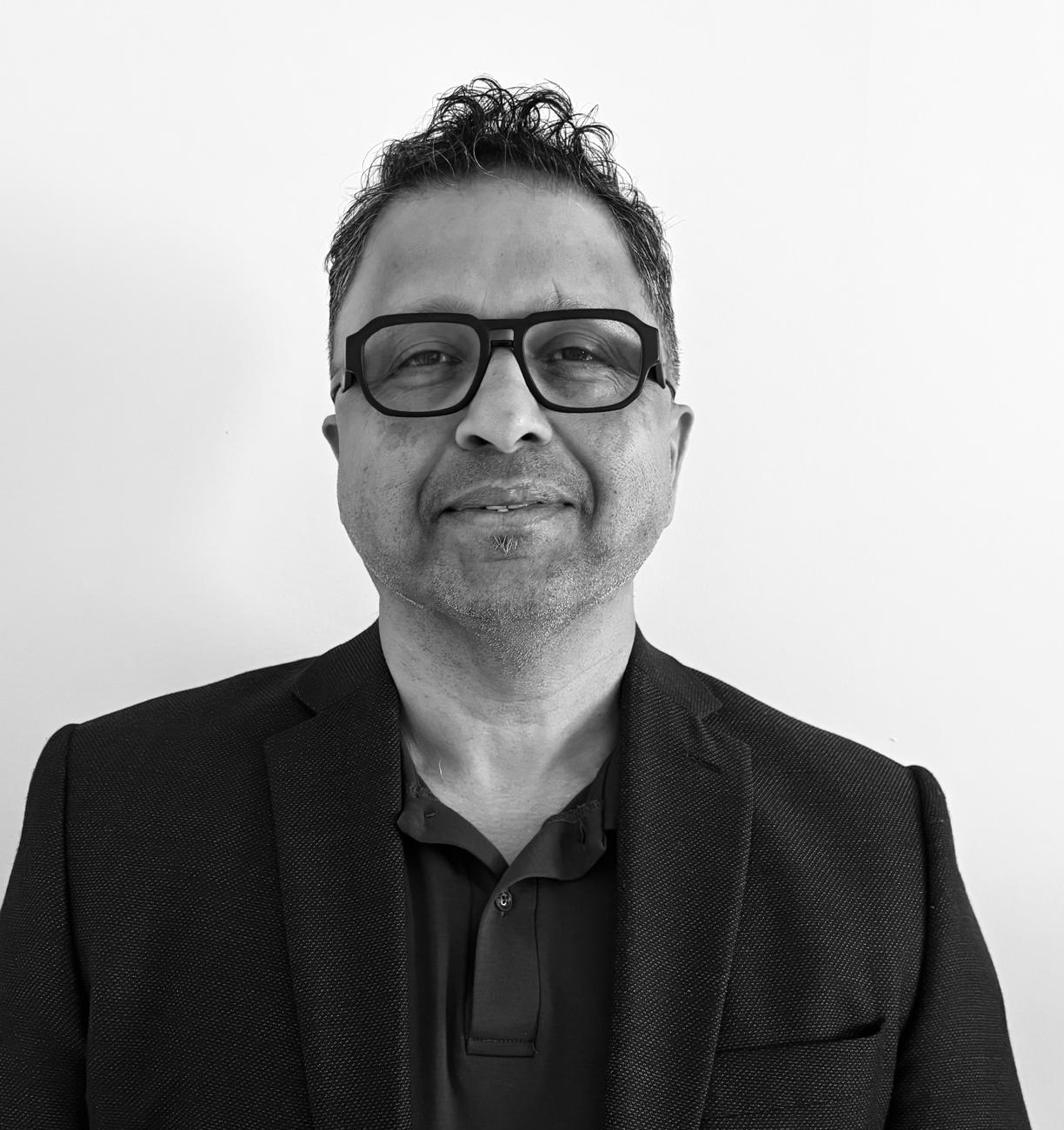 A man wearing glasses, a dark blazer, and a polo shirt stands against a plain white background, looking at the camera with a neutral expression.