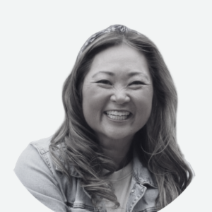 A woman with long hair smiles broadly, wearing a denim jacket and a headband, against a plain white background.