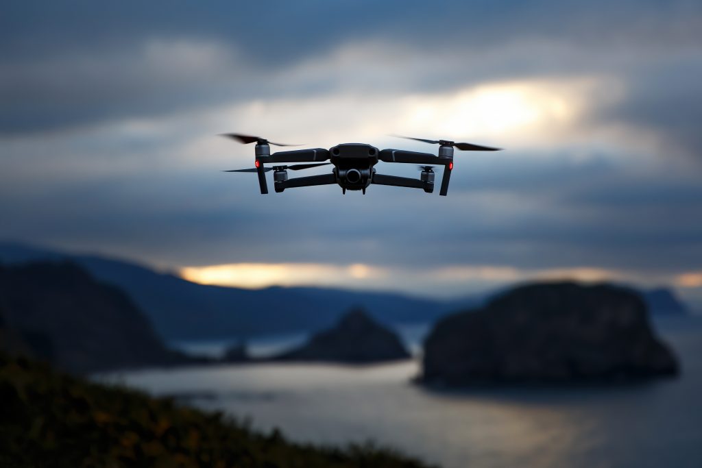 A drone hovers in midair at dusk above a coastal landscape with cliffs and water in the background.