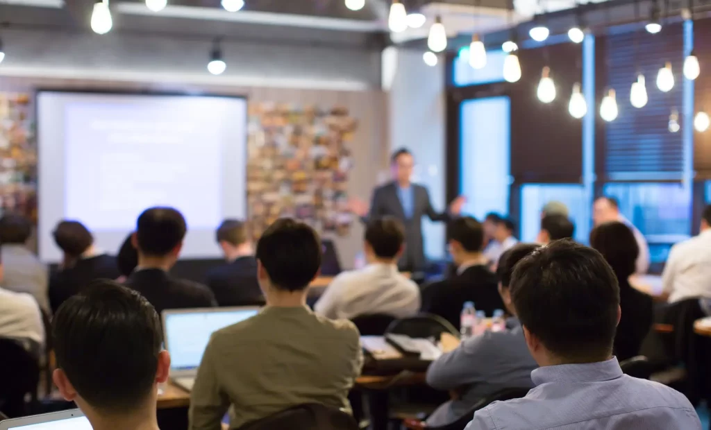 A group of people sits facing a speaker giving a presentation in a warmly lit room. There is a projector screen displaying a slide in the background.
