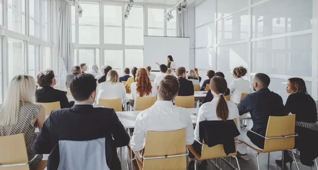 A group of people seated in a well-lit room listening to a speaker at the front, who is presenting information on a screen.