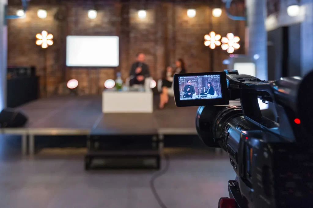 A video camera records a live event on a stage, where two people sit in front of a projected screen with blurred visuals and lights in the background.