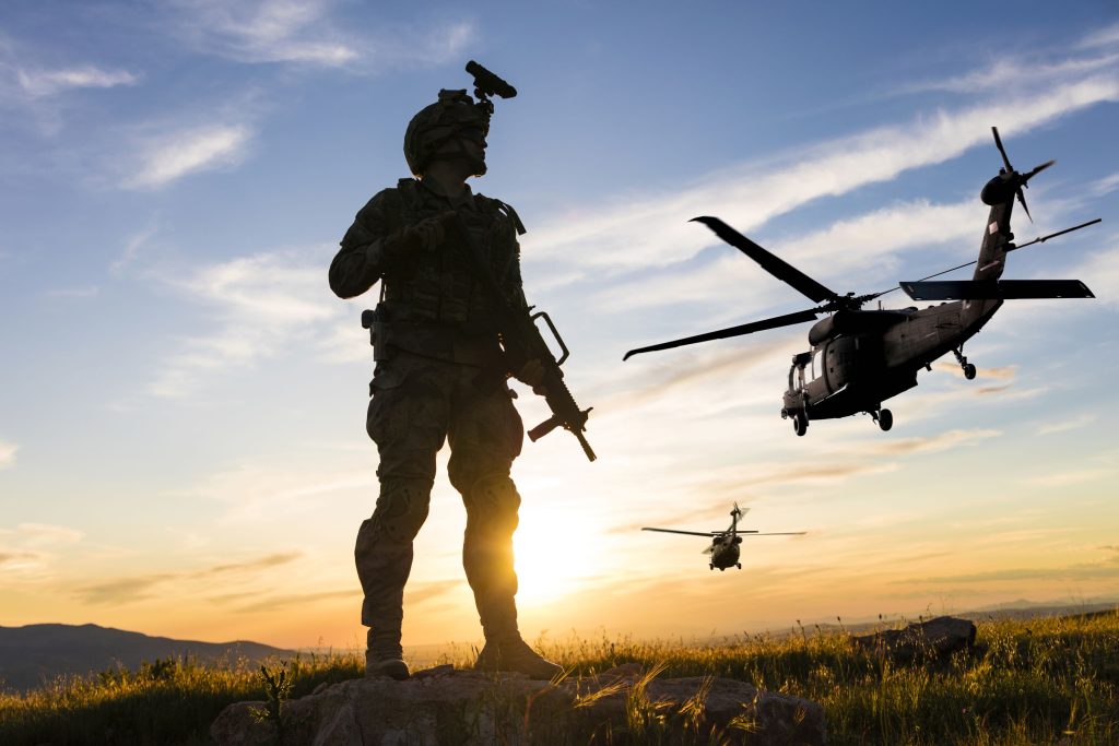 A silhouetted soldier standing with a rifle in hand. The sun is rising on the horizon and two helicopters are flying overhead. 