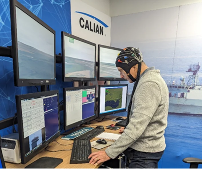 A person wearing a headset stands at a workstation with multiple monitors displaying various data and images, including maritime scenes. The room has a blue theme with the word "CALIAN" on the wall.