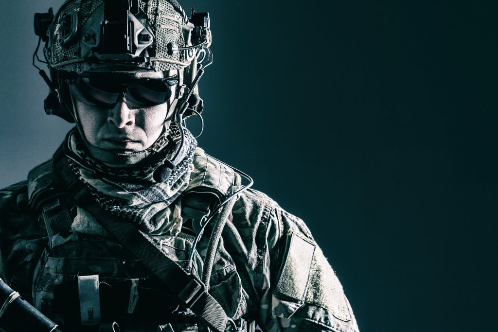 A soldier in full tactical gear, including a helmet, sunglasses, and camouflage uniform, stands against a dark background.