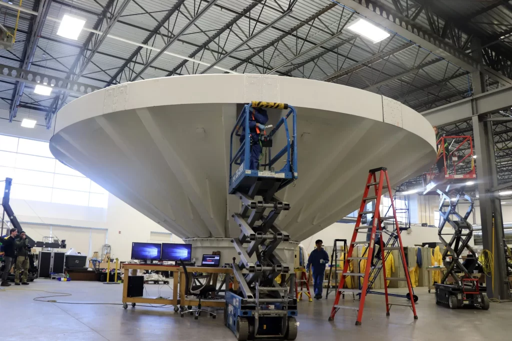 A large, white, conical structure inside a spacious industrial facility, surrounded by scaffolding, ladders, and workers. Multiple workstations with computers and other equipment are visible.