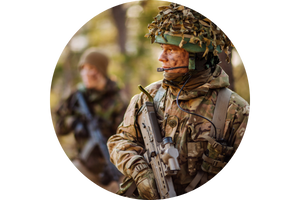 Two soldiers in camouflage uniforms and helmets, one in the foreground with a rifle and communication gear, the other slightly blurred in the background, standing in a wooded area.