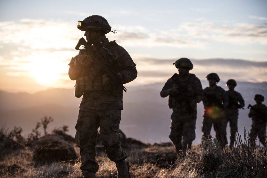 A group of soldiers in full combat gear, including helmets and rifles, walk through a rugged landscape during sunrise.