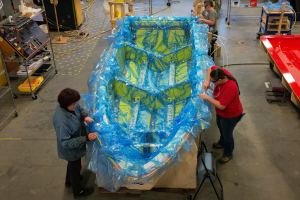 Workers cover a boat with a blue and yellow plastic sheet in a workshop.