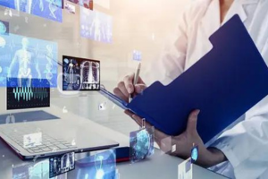 A person in a lab coat holds a blue folder and writes on a document while standing in front of a laptop displaying medical images and data visualizations.