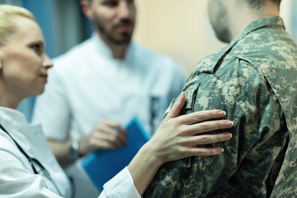 Two doctors speak with a person in military uniform; one doctor places a reassuring hand on the soldier's shoulder.