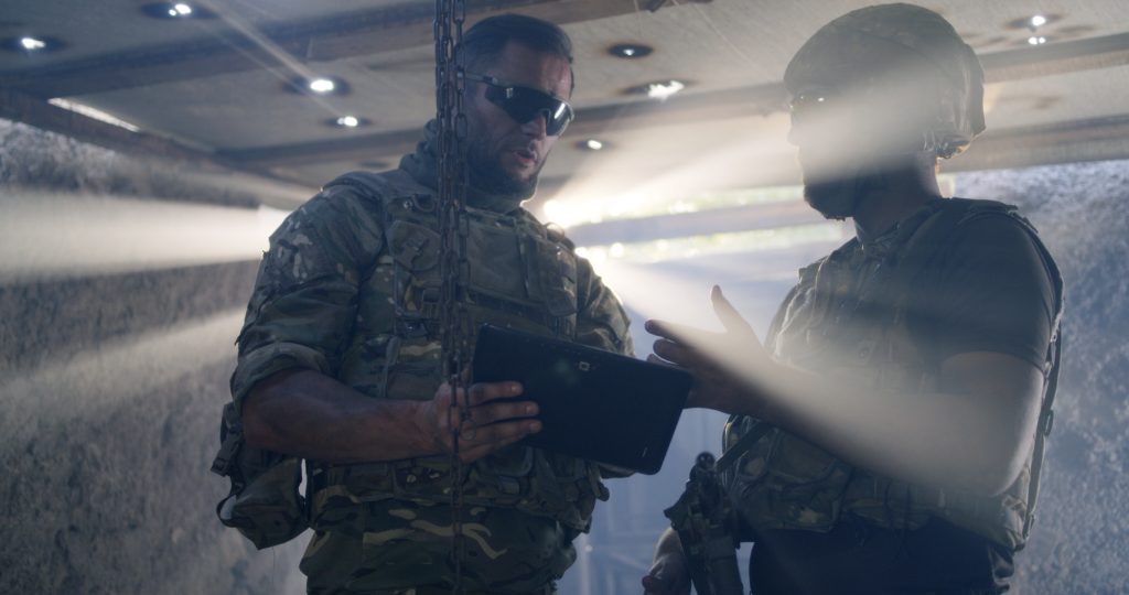 Two soldiers in camouflage uniforms and tactical gear converse while looking at a tablet in a dimly lit area with beams of light coming through openings above.