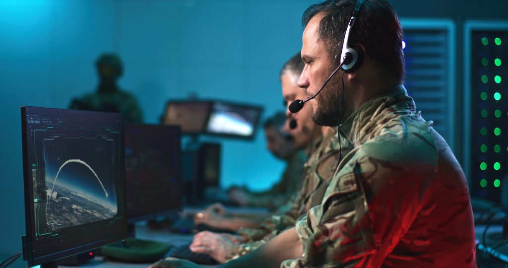 Military personnel in camouflage uniforms monitor and operate computer systems with headsets in a control room environment.