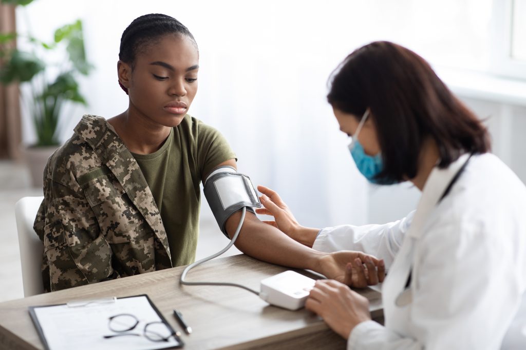 A healthcare professional checks the blood pressure of a person in military attire sitting at a desk.