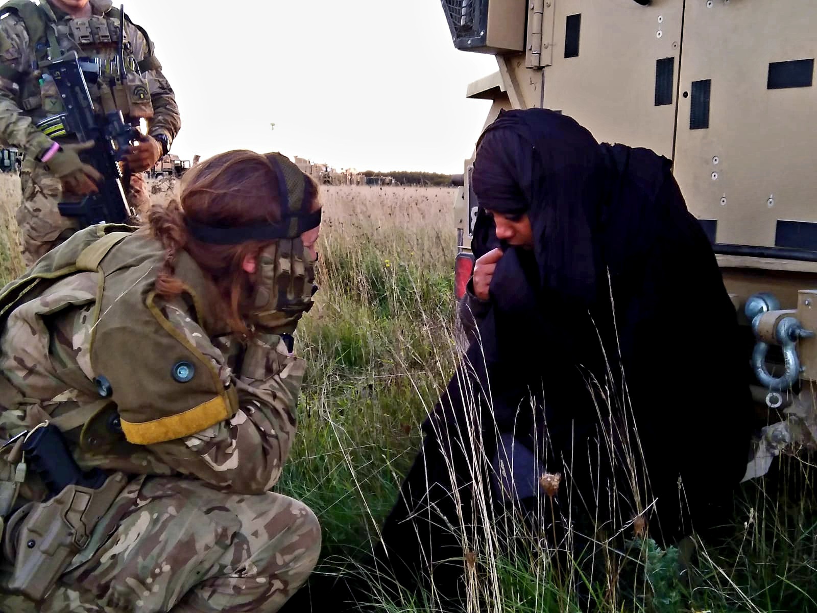 A woman in a black headscarf crouches beside a person in military gear in a grassy field.