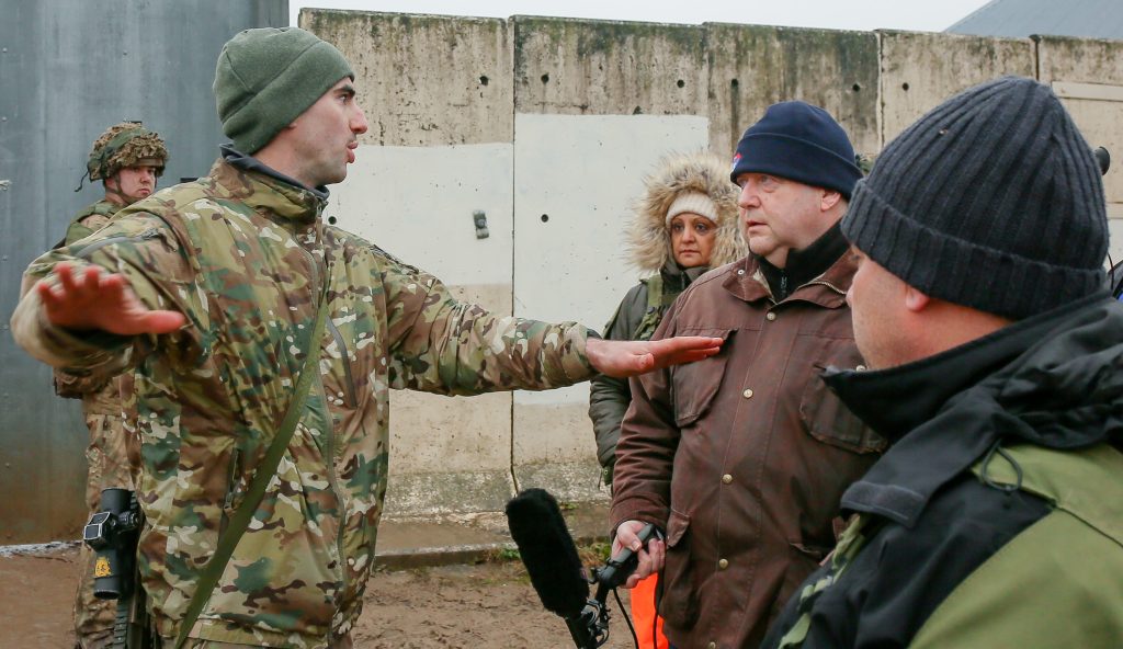 A person in military attire gestures while speaking to a group of people dressed in winter clothing, standing near a concrete wall. Two other military personnel stand in the background.