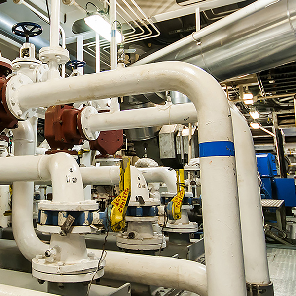 Industrial setting with white pipes, valves, and yellow tags in a mechanical room. Overhead lighting illuminates the intricate network of ducts and equipment.