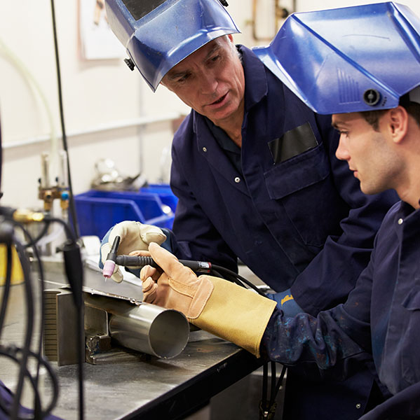 Two people in protective gear work with welding equipment at a metalwork station.