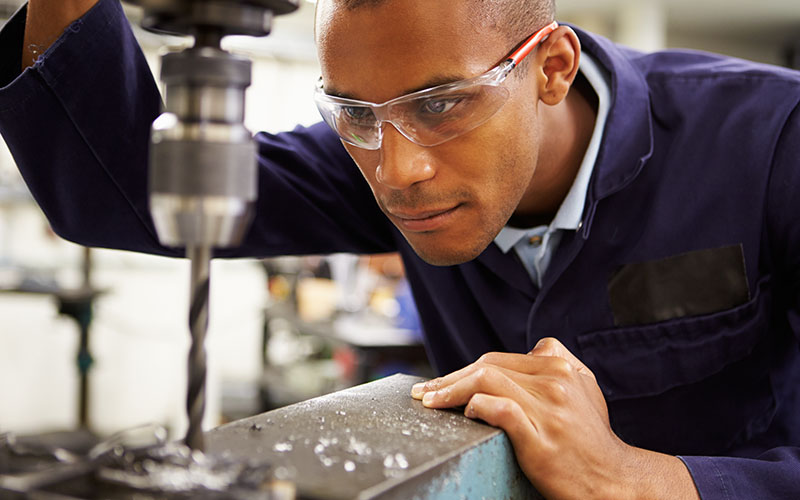 A person wearing safety goggles operates a drill press in a workshop.