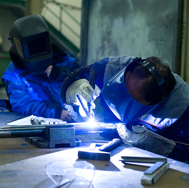 Two people wearing protective gear are welding metal parts on a workbench. One is actively welding, and the other is observing.