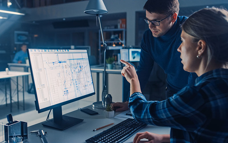 Two people in an office look at a computer screen displaying technical schematics. One person points at the screen while the other stands beside them.
