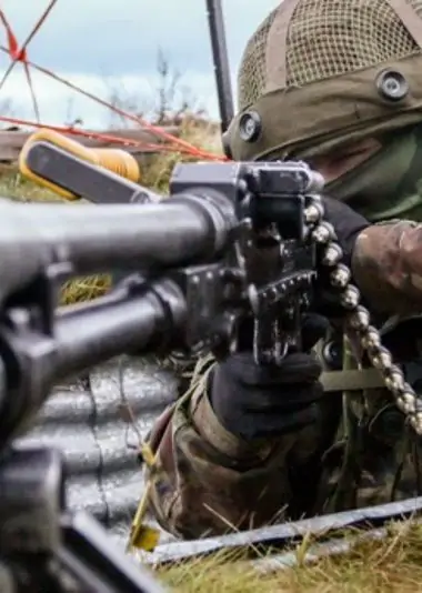 A soldier in camouflage and helmet aims a large weapon, lying prone in a grassy area with metal and wires in the background.