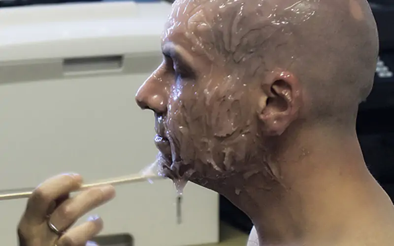 Person with a bald head getting a translucent facial mask applied with a stick.