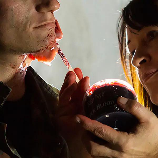 Makeup artist applies fake blood to an actor's chin using a brush, holding a jar labeled "Thick Blood.