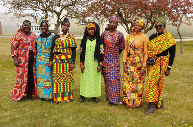 Seven people standing outdoors, wearing brightly colored traditional African clothing, with trees and a building in the background.