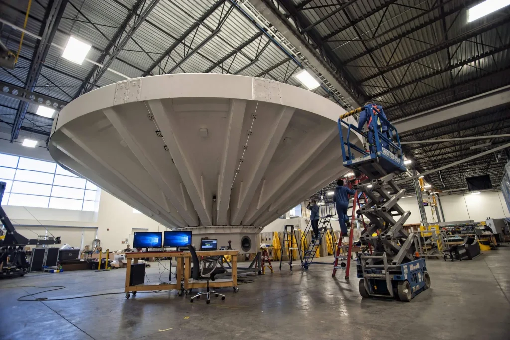 Large white satellite dish under construction inside an industrial facility, with workers on a scissor lift and equipment scattered around.