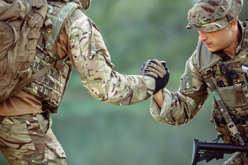 Two soldiers in camouflage uniforms clasp hands, one helping the other up. They are outdoors, and military gear is visible.