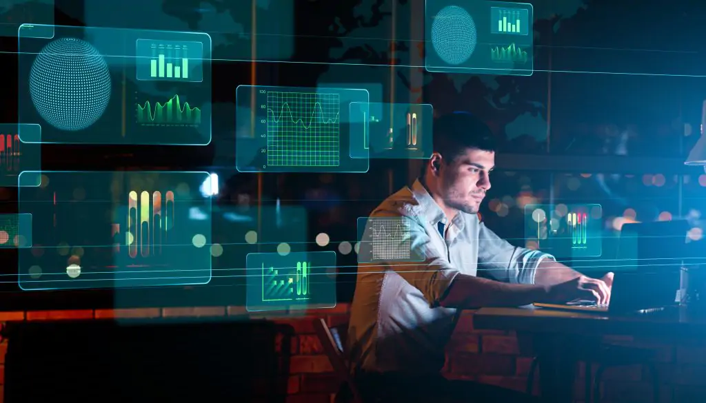 A man sits at a desk using a laptop, surrounded by digital data graphs and charts projected in the air, suggesting data analysis or digital work.