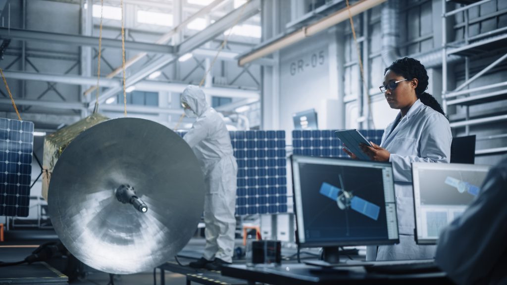 Engineers in a laboratory inspect satellite equipment and solar panels; one person uses a tablet while others work on machinery, with computer monitors displaying satellite images.