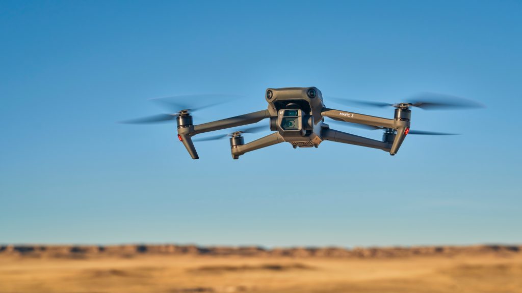 A black quadcopter drone with a camera hovers in the air against a clear blue sky above a blurred, arid landscape.