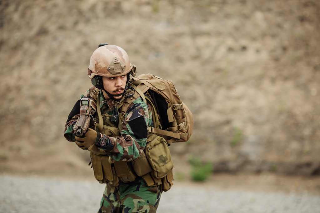 A soldier in camouflage uniform, helmet, and backpack holds a rifle in a ready position outdoors on rocky terrain.